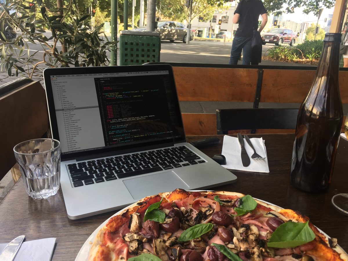 A computer showing code at the back of a table with a pizza in front of it
