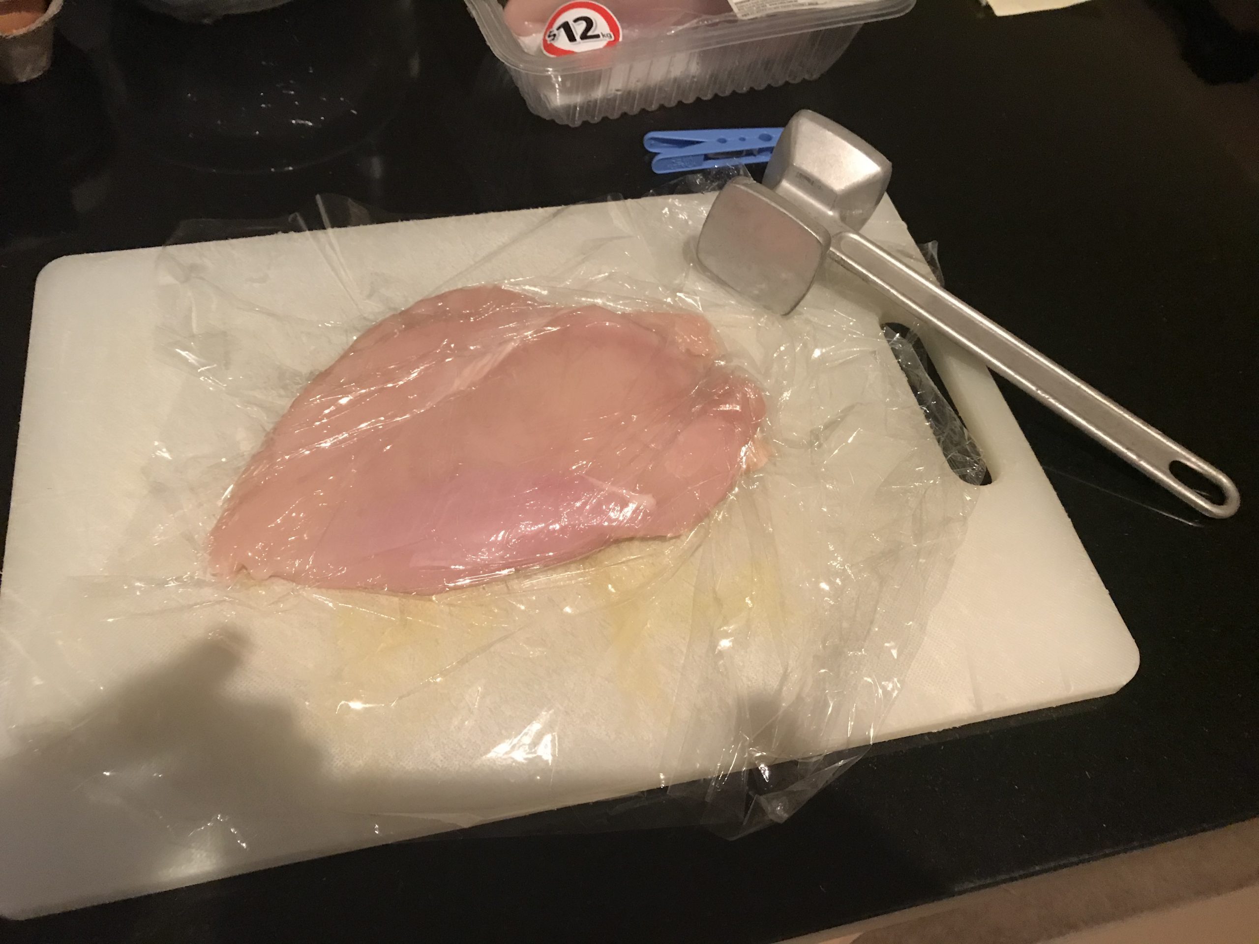 Uncooked chicken breast under cling wrap with a tenderizing mallet sitting next to it
