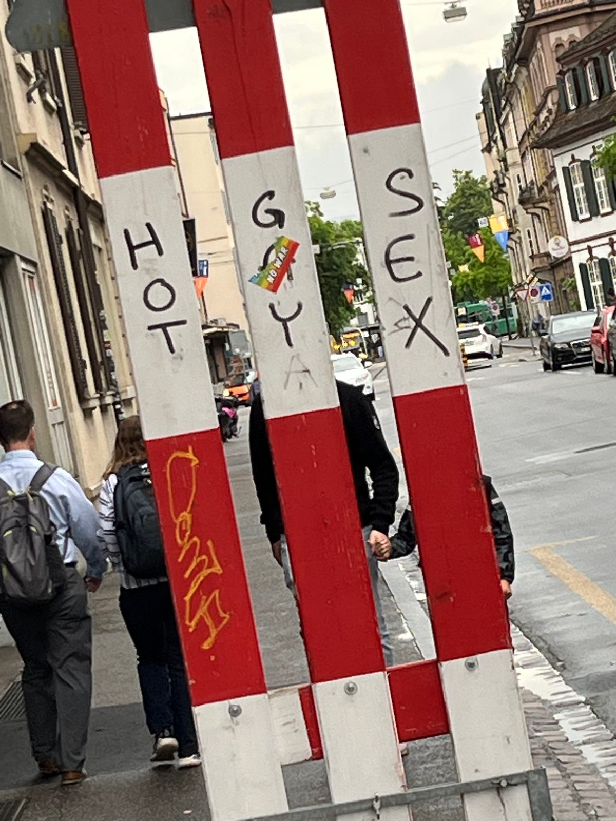 A warning sign made up of three vertical planks. The sign is painted in red and white horizontal stripes. In a white section HOT GAY SEX is graffiti on the sign.