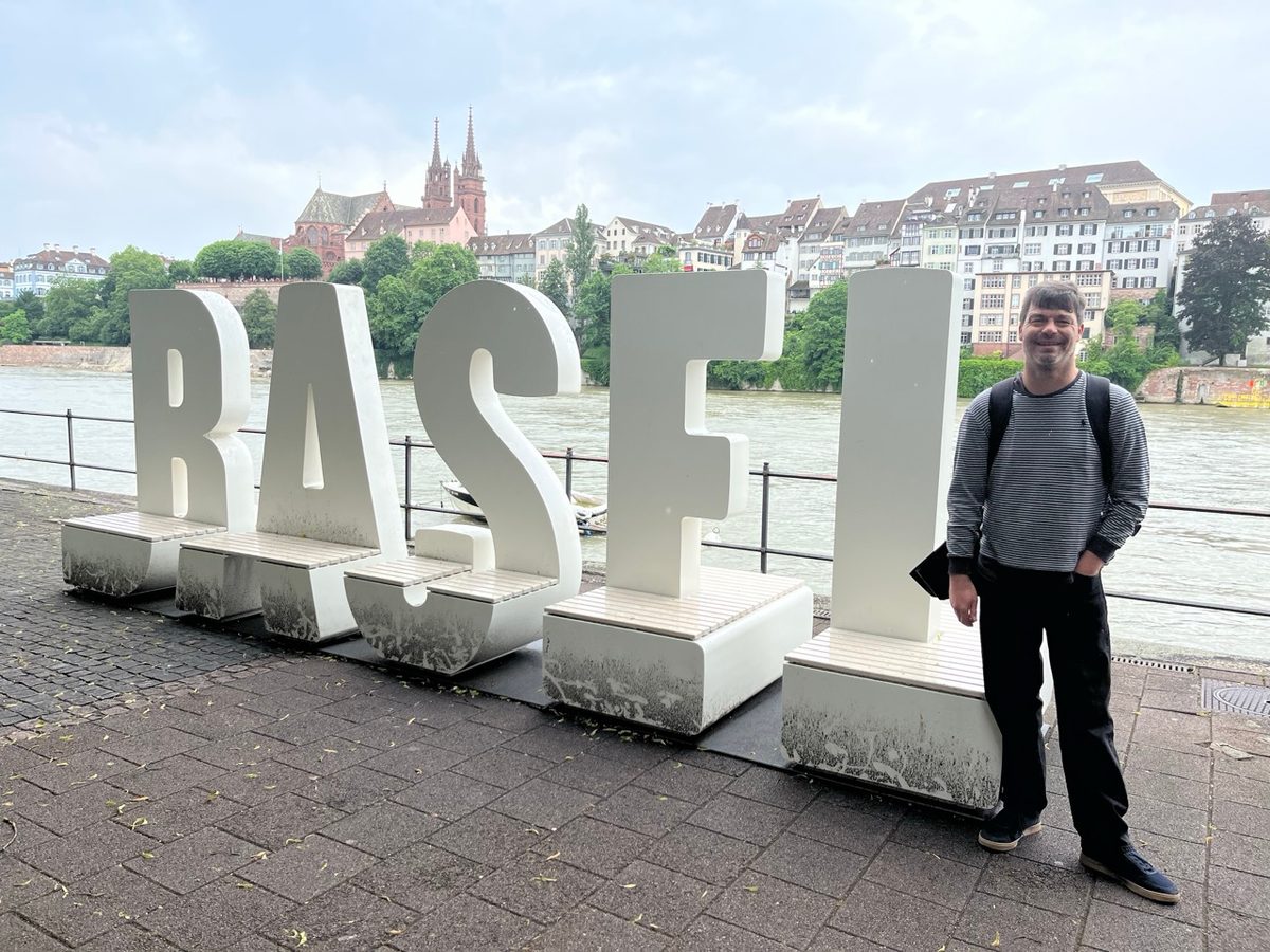 Me standing next to a six foot high white sign that says Basel with the Rhine and the old city in the background.