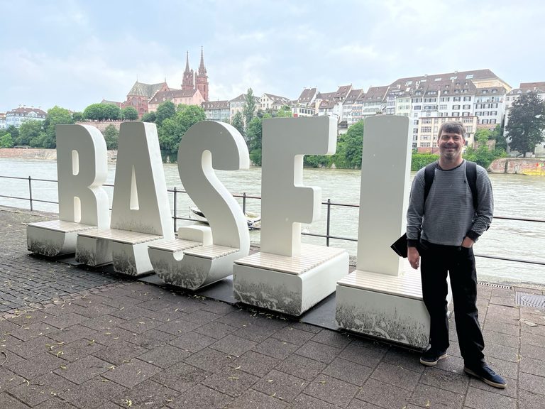Me standing next to a six foot high white sign that says Basel with the Rhine and the old city in the background.